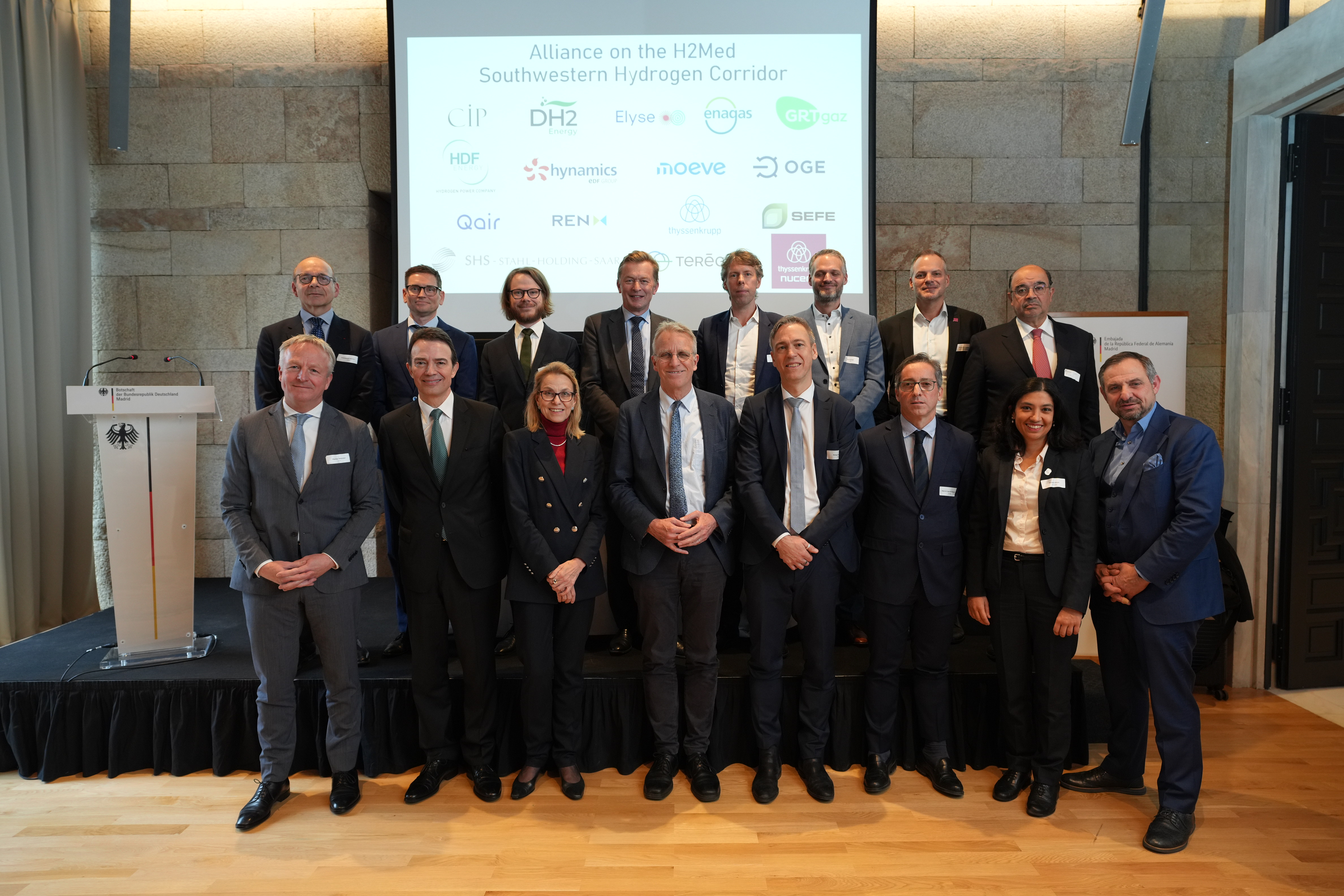 Sechzehn Personen stehen vor einem Podium und schauen in die Kamera.